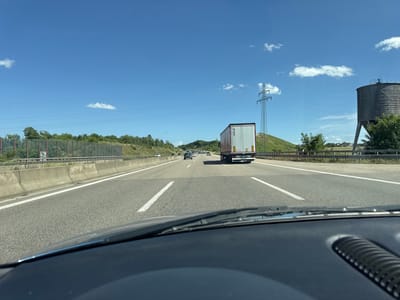 The image shows a view from the inside of a car traveling on a highway. The highway has multiple lanes and is bordered by concrete barriers and green hills with trees. A large semi-trailer truck is visible in the right lane, ahead of the car. The sky is blue with scattered white clouds, and a tall electrical pylon is visible on the right side of the road. In the distance, more cars and trucks can be seen on the highway. A large, cylindrical silo structure is also present on the right side of the highway, behind the electrical pylon. The car's dashboard and windshield wipers are in the foreground. This scene is located on the A8 Autobahn near Wimsheim, Germany.