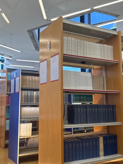 The image shows a section of bookshelves, likely in a library. The shelves are made of light-colored wood. The books on the top shelves are stacked tightly, most with white spines and some with dark blue spines. Some of the spines have text printed on them. Two white rectangular papers are affixed to the underside of the top shelf, and a small metallic plate with the number "2" is also visible. The angle of the image suggests it was taken from below, looking up at the shelves. Light fixtures are visible on the left side of the image. A window lets in natural light. Additional shelves are visible below. The exact location in Sant Vicent del Raspeig, Spain is indeterminable from the image.