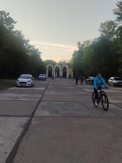 The image depicts a scene in a park or public square in Zhovti Vody, Ukraine. The foreground features a paved pathway with distinct concrete slabs and a dark line running between them. A woman wearing a blue jacket and black pants is cycling on a black bicycle, moving from left to right across the pathway. In the middle ground, there are several parked cars, including a white hatchback on the left and a dark SUV. Further in the background, a large architectural structure with multiple arches, painted yellow and white, is visible. Several people are standing near this structure. The background is dominated by lush green trees and a pale sky with some faint clouds. The lighting suggests it is late afternoon or early evening.