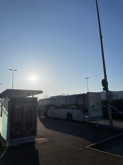 The scene depicts an outdoor urban environment under a clear, bright blue sky with the sun low on the horizon, casting long shadows across an asphalt surface. The location is the city of Plaisir, France, as indicated by "VILLE PLAISIR" text on an advertising panel.

In the foreground, a modern bus shelter with a glass panel is positioned on the left, containing an illuminated advertisement for "86 CREERY." To its right, a light-colored passenger bus, branded "Île-de-France Mobilités" and identifiable as a SETRA model, is stationary, oriented towards the right. A second, darker bus is partially visible behind the first.

Further to the right, a tall, dark utility pole stands prominently, adorned with a circular red and white 'no entry' or 'stop' sign. Adjacent to this pole is a freestanding advertising panel displaying a map or route information under the heading "VILLE PLAISIR." A dark-colored vehicle is partially visible behind this pole and panel.

Two streetlights with twin lamp heads are situated on the left side of the frame. Bare trees are visible in the background between the buses and some distant industrial or commercial buildings. No individuals are visible in the frame.