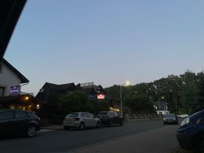 This image captures a quiet street scene in Bolesławiec, Poland, likely at dusk. The sky is a pale blue, transitioning into a darker hue as night approaches, suggesting early evening. The overall atmosphere is calm and peaceful.

The scene is dominated by a street lined with buildings and trees. On the left, a building with a stone facade features a prominent sign illuminated in purple with the word "RECPCJA" in white, indicating a reception area. Adjacent to this, a dark-colored building with a wooden exterior appears to be a restaurant or hotel, also with illuminated signs. One sign, colored red and white, reads "RESTAURACJA."

Several cars are parked along the street and on the roadside. A dark grey hatchback is parked on the left, and a light-colored hatchback is parked further down on the right. A black SUV is parked in front of the "RESTAURACJA" building, and another dark-colored vehicle is visible in the distance. A partially visible blue vehicle is in the foreground on the right.

The background is filled with dense green trees, suggesting a natural, possibly forested, setting surrounding the buildings. A streetlamp casts a warm yellow glow on the street, illuminating the area. The architecture of the buildings, particularly the dark wooden structures with pitched roofs, hints at a European style. The presence of both a "RECPCJA" and "RESTAURACJA" sign suggests this is an establishment that offers accommodation and dining services. The moon is faintly visible in the sky.