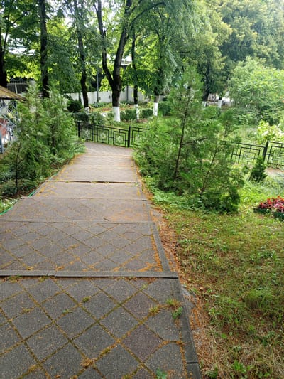 The image shows a paved pathway with a diamond pattern leading down a gentle slope in a park-like setting. The pathway is bordered by grass on the right and a row of trees and bushes on the left. A black metal fence runs along the right side of the pathway. In the background, there are more trees, some with white-painted trunks, and a white wall. A small flower bed with red flowers is visible to the right of the pathway. The scene appears to be in the city of Giaginskaya, Russia, given the context provided. There are no people visible in the image. The environment is green and appears to be a public outdoor space.