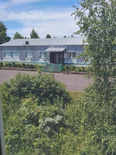 The image shows a one-story, rectangular building with a corrugated metal roof and walls. It has several windows with green trim and an entrance with a covered porch and green steps. The building appears to be in a rural or semi-rural setting, with trees and vegetation surrounding it. A gravel path leads to the entrance. In the foreground, there is dense green foliage. The sky is blue with white clouds. The image was taken in Новый Каракан, Russia.