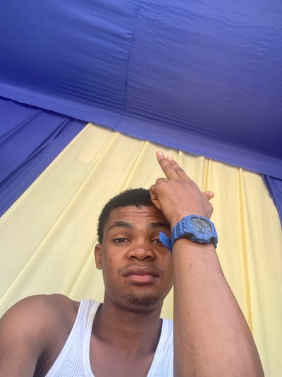 A close-up image depicts a male individual, appearing to be a young adult, looking directly at the camera. He has short, dark, curly hair and light stubble on his chin. He is wearing a white mesh tank top. His right arm (viewer's left) is bent, with his right hand resting on top of his head, fingers slightly splayed. A blue digital wristwatch with a robust casing is worn on his right wrist. The background consists of alternating vertical panels of royal blue and light yellow fabric, suggesting an indoor setting with decorative drapes, possibly within a tent or an event space. This scene is located in Ikot Ete Ekpo, Nigeria.