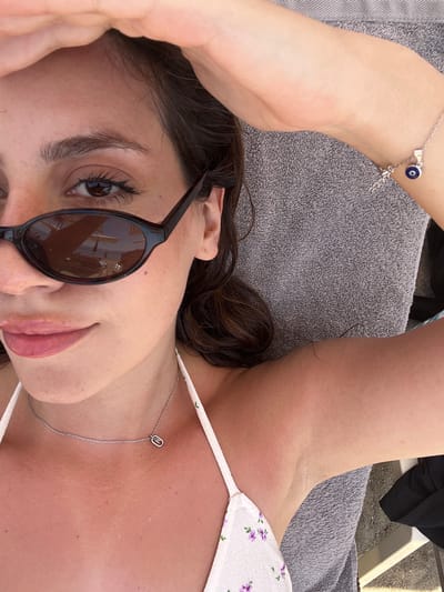 A close-up shot captures a woman from the chest up, lying on a grey towel, presumably in Troulos, Greece. Her right arm is raised, shielding her forehead from the sun with her hand. She wears sunglasses with dark lenses, reflecting a beach scene with umbrellas and people. A delicate silver necklace with a rectangular pendant rests on her collarbone. A white bikini top with a subtle floral pattern is visible. On her left wrist, a silver bracelet adorned with a blue and silver evil eye charm is worn. The lighting suggests it is daytime, with a warm, natural light illuminating the scene. The background is out of focus, but hints at an outdoor setting, likely a beach resort given the sunglasses reflection and the woman's attire.