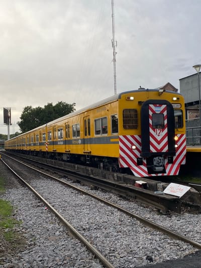 An image captures a bright yellow and black train from a side-front perspective, positioned on railway tracks under an overcast sky. The train appears to be stationary, possibly at a station or siding.
The front section of the train features a striking black panel with bold red and white chevron stripes, which serve as a high-visibility marking. A small sticker of the Argentine flag is visible above the main windshield, and below it, the number "17" is displayed in red on a white background, accompanied by a red chevron pointing to the right. Two rectangular headlights are illuminated. Above the windshield, the text "GRAL. LEMOS" is visible on the yellow body.
Along the side, the train car is primarily yellow with horizontal grey stripes, featuring multiple windows. The words "Metrovías" and "Línea Urquiza" are clearly visible, identifying the operator and the specific railway line. The car number "M.U. 3900" is also legible.
Two parallel railway tracks, covered with grey gravel ballast, extend from the foreground into the distance. Adjacent to the tracks, a white warning sign on a post prominently displays "PELIGRO" (Spanish for "DANGER") in red uppercase letters, with additional smaller text below.
The sky is uniformly overcast and light grey, suggesting a cloudy daytime. In the background, power lines, parts of buildings, and green tree foliage are discernible. The scene depicts a typical railway environment, consistent with a location such as Sáenz Peña, Argentina. No people are visible in the image.