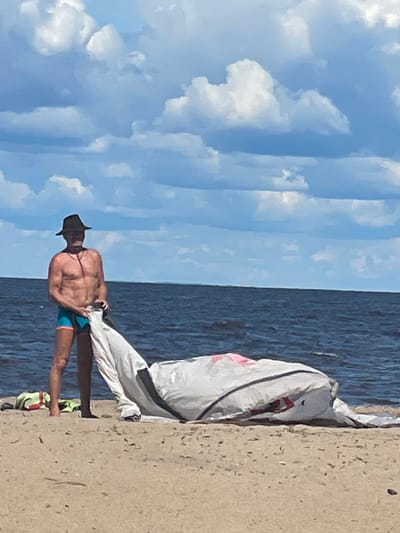 A shirtless man wearing a wide-brimmed hat and swim trunks stands on a sandy beach. He is holding a white and gray object, possibly a kite or sail, which is spread out on the sand in front of him. The man is looking down at the object. The ocean is in the background, with a blue sky and clouds above. The location is the beach in Belozersk, Russia.