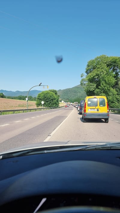 The image shows a view from inside a car on a sunny day, looking out at a traffic jam on a two-lane road. The road is asphalt, with white lane markings. On the left side of the road, there is a guardrail, a traffic light pole with a solar panel, and a large green tree. Beyond the guardrail, there are fields and hills in the distance. On the right side of the road, there are several cars, including a yellow van with the license plate "1234 DGX". The traffic is backed up, indicating a slowdown. The sky is clear and blue. The overall scene suggests a typical day with road congestion, likely on a route leading to or from Olot, Spain, given the presence of rolling hills and green vegetation characteristic of the region.