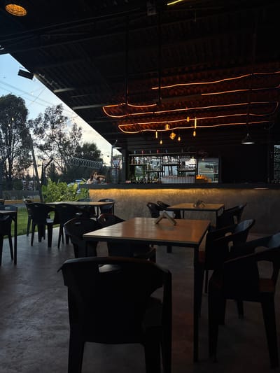 The image depicts a dimly lit outdoor seating area of a restaurant or bar, likely in San Pedro Cholula, Mexico. Rows of black plastic chairs are positioned around square wooden tables. String lights hang above, creating a warm glow. Through an opening, a view of the outside is visible, including trees, a fence, and the sky. In the distance, people are seated at tables outside the fenced in area, and they appear to be engaging in conversation. The scene is captured from a slightly elevated perspective.