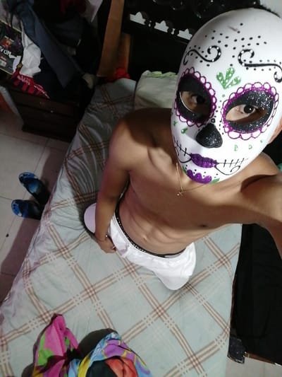 A shirtless person wearing a white Day of the Dead mask is sitting on a bed with a light blue and brown plaid comforter. The person is wearing white underwear, a gold necklace, and a thin bracelet. Blue sandals are visible on the floor. The background shows a cluttered room with a wooden dresser, clothes, and other personal items. A colorful cloth is visible at the bottom left of the image.