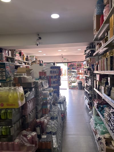 The image shows the interior of a retail store, likely a supermarket or grocery store, located in Mouscron, Belgium. Shelves stocked with various goods, including bottled water, canned drinks, and other packaged food products, line the aisles. The store appears to be well-lit, with a tiled floor and recessed lighting in the ceiling. The perspective is looking down one of the aisles towards the entrance of the store, with shelves extending along either side.
