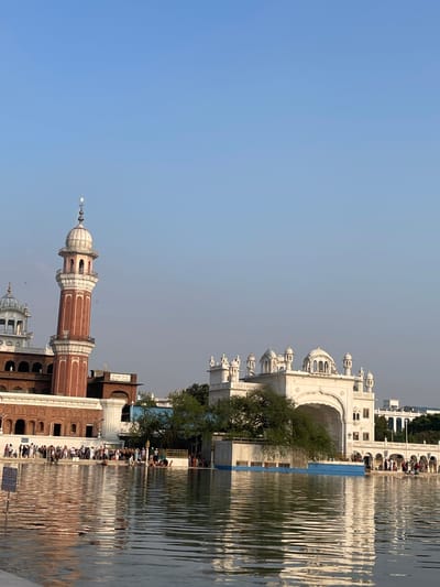 The image depicts the Golden Temple complex in Amritsar, India, seen across a large body of water. The foreground is dominated by the rippling surface of the water, reflecting the sky and the structures on the far side.
On the left, a tall, octagonal minaret with red brick and white decorative elements rises against a clear blue sky. It is topped with a white dome and spire. Adjacent to it, a building with a similar architectural style, featuring arched windows and a domed roof, is partially visible.
In the center and right of the image, a series of white, domed structures with intricate architectural details are prominent. These buildings appear to be part of a larger complex, characterized by arches, pillars, and multiple smaller domes. A large, arched entryway is a central feature of the white structures.
A tree with lush green foliage is situated in front of the white buildings, partially obscuring them. Along the edge of the water, a multitude of people are gathered, some standing, some sitting, and some appearing to be bathing or washing. They are dressed in various traditional and contemporary clothing.
The environment suggests a bright, possibly late afternoon or early morning, with clear skies and soft lighting illuminating the scene. The overall atmosphere is one of reverence and activity surrounding a significant religious site.