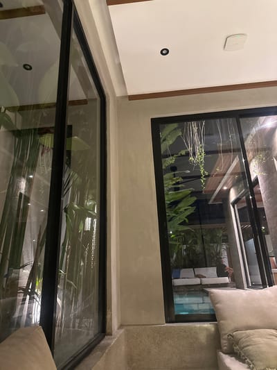 The image shows an interior view, possibly in a residential or commercial building in Tumbak Bayuh, Indonesia. A floor-to-ceiling window offers a view of lush greenery and possibly other structures outside. The window's reflection reveals interior design details such as furniture and architectural elements. The interior features a white wall with a mounted device and a lighting fixture. The overall lighting suggests an evening or nighttime setting.