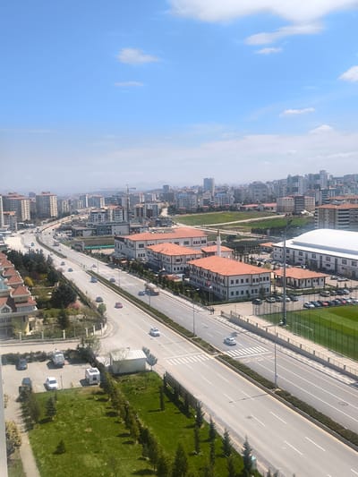 This media file captures an elevated view of an expansive urban landscape in Etimesgut, Turkey, on a bright, sunny day. The sky is a clear blue with scattered white clouds, and the scene is illuminated by strong daylight.

A prominent, multi-lane road curves through the center of the image, bustling with various cars, trucks, and a white camper trailer visible in a nearby parking lot. Several pedestrians can be seen walking along the sidewalks. The road features clear white lane markings and pedestrian crosswalks.

The urban setting comprises a mix of architectural styles. To the right of the main road, a complex of buildings with distinctive red-tiled roofs stands out, one of which features a small minaret and a Turkish flag flying nearby, suggesting a mosque or a culturally significant institution. Adjacent to this complex is a large, modern building with a white roof and a well-maintained green football field surrounded by a fence, indicating a sports or educational facility. Nearby, parking lots are filled with numerous vehicles.

Further into the city, multi-story residential and commercial buildings dominate the skyline, some with visible text, such as "Doga" on a commercial facade. Lush green areas, trees, and smaller residential structures with their own green spaces line the roads. In the distant background, hazy mountains frame the horizon, and construction cranes are visible among the urban sprawl. The overall scene depicts a vibrant, busy city under pleasant weather conditions.