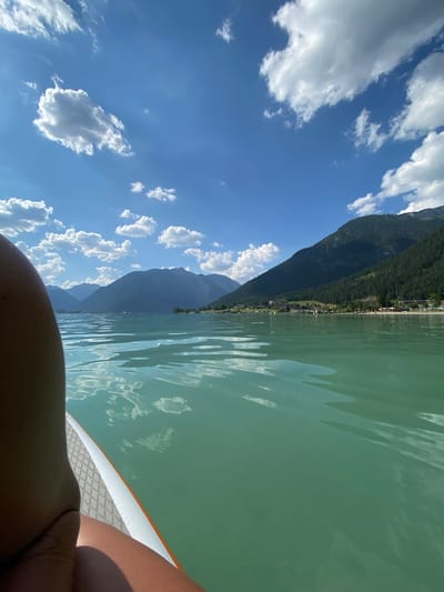 This media file captures a serene moment on a calm, turquoise lake, likely during daytime in fair weather, given the bright sunshine and scattered white clouds against a vibrant blue sky. The scene is dominated by a large, tree-covered mountain range that slopes down towards the water's edge, where a small village with scattered buildings is nestled. The perspective is from someone on a paddleboard, looking out over the tranquil water. Reflections of the sky and clouds are visible on the water's surface, creating a rippling, abstract pattern. The foreground is partially occupied by the legs and torso of the person on the paddleboard, suggesting they are in a relaxed, reclined position, enjoying the view. The overall impression is one of peaceful immersion in nature, with the vastness of the sky and mountains providing a sense of calm and adventure. The location is identified as Lärchenwiese, Austria, a detail that adds context to the mountainous and lake landscape. There is no visible text in the media file.