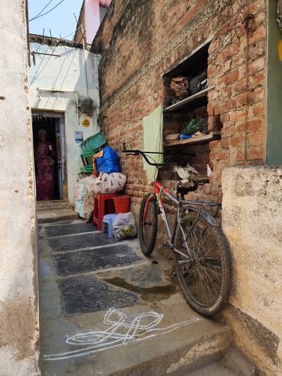 This image depicts a narrow outdoor passage in Pedda Kammavaripalli, India, bordered by two buildings under a clear blue sky. The left wall is light-colored and textured, with an open wooden doorway revealing an elderly woman. She has white hair, wears a red and pink patterned saree, and holds a white cup in her right hand. An electrical meter, white piping, and utility signs ("HAC 5008" and "159") are mounted on the wall near her doorway. Stacked against the wall are various items, including a blue bag, a white floral-patterned bag, a white sack, a red plastic stool, a blue plastic stool, two orange plastic buckets, and several green plastic baskets.
The right wall is constructed of exposed reddish-brown brick, featuring two recessed shelf-like niches. These niches contain shoes, bags, and miscellaneous items, with a light green cloth draped from the lower shelf. Leaning against this brick wall is a red and grey mountain-style bicycle with a black seat, black tires, and a rear fender. The ground consists of dark grey stone slabs interspersed with lighter concrete, displaying several white chalk geometric patterns (kolam/rangoli) on the lower steps and sections of dampness. Overhead, utility wires are visible against the bright sky.