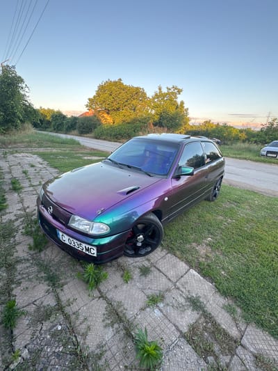 The image shows a custom purple and green iridescent car parked on a cracked concrete path with grass growing through it, bordering a dirt road. The car is a hatchback with black rims and a prominent license plate that reads "C 0335 MC". In the background, there are trees, a building with a red roof, and other vehicles on the road. The sky is clear blue with a hint of sunset colors on the horizon. The overall scene suggests a rural or semi-rural setting, likely in or near Lepitsa, Bulgaria, given the license plate format and the presence of a structure that could be a house.