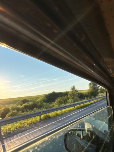 The image is a side view from inside a vehicle, looking out the passenger window. The scene depicts a road with a guardrail running alongside it, bordered by a green, grassy field with scattered trees and bushes. The sky above is a clear blue with wispy white clouds, and the sun appears to be low on the horizon, casting a warm, golden light on the landscape. The interior of the vehicle is visible in the foreground, with the dark frame of the window and the reflective surface of the side mirror. There are no people or specific actions captured in the image. The location is identified as Verkh-Sosnova, Russia.