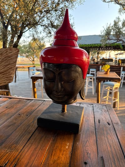 A close-up, eye-level shot captures a dark-faced Buddha head statue with a bright red, pointed headdress, displayed on a weathered wooden table. The statue is the central subject, with its closed eyes and serene expression. Behind the statue, an outdoor seating area of a establishment is visible, featuring wooden tables and white stools. Olive trees with dappled sunlight filtering through their leaves frame the scene. In the background, a building with a covered walkway and further seating areas are discernible, suggesting a relaxed, possibly coastal, environment. The setting is identified as Angelochori, Greece, during what appears to be late afternoon or early evening, given the warm, golden light illuminating the wooden surfaces and casting soft shadows. No people are visible in the frame.