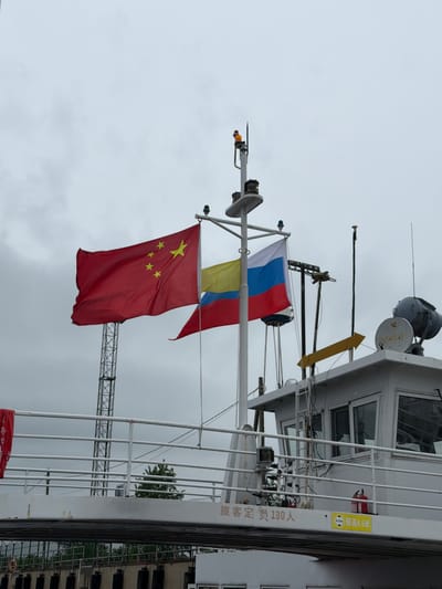 The image shows a portion of a white boat or ship set against an overcast, grey sky. Two national flags are prominently displayed on a mast: the Russian flag (white, blue, and red horizontal stripes) flies above the Chinese flag (red with a large yellow star and four smaller yellow stars). Both flags appear slightly damp, suggesting recent rain or mist.

The upper deck or bridge structure of the boat is visible on the right side of the frame. It features multiple windows, white railings, and various pieces of equipment, including antennae and satellite dishes.

Chinese text is visible on the side of the boat: "旅客定员 130人," which translates to "Passenger capacity 130 people." Above this, a yellow rectangular sign shows a red fire extinguisher icon and the characters "最高4.5米," indicating "Maximum height 4.5 meters."

In the background, a metal truss structure, likely part of a port crane or gantry, is partially visible, along with some green foliage from a tree. The muted lighting suggests an overcast daytime. The presence of both Russian and Chinese flags on a vessel, alongside Chinese text, strongly suggests an international connection, aligning with the context of Nizhneleninskoe, Russia, which is a border town with China on the Amur River.