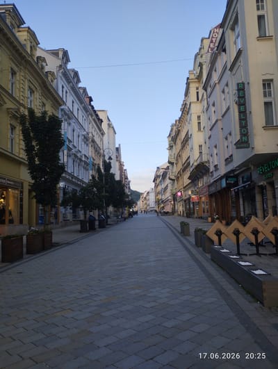 This image shows a pedestrian street lined with buildings on both sides. The buildings are several stories tall and appear to be hotels or businesses with shops on the ground floor. Several trees are planted along the sidewalk, and there are planters with flowers. In the foreground, there are outdoor cafe tables set up. The street is paved with cobblestones. The sky is clear and a light blue, suggesting it is either early morning or late evening. A timestamp at the bottom right corner indicates the date and time as 17.06.2026 20:25. Signs visible on the buildings include "HOTEL", "RÉVELTON SUITES", "BAGEL LOUNGE", and "Fashion PANSKA MOD". There are a few people walking in the distance, but the street is not crowded. The overall scene evokes a sense of tranquility and a pleasant evening stroll in a European town.
