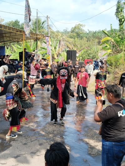 In the city of Kerjo, Indonesia, a traditional performance is underway outdoors. The ground is wet, reflecting the light, suggesting recent rain or a deliberate wetting of the performance area. Several individuals are dressed in elaborate costumes, some wearing masks that resemble stylized demonic faces with prominent teeth and red accents. One performer in the foreground wears a black costume with a red scarf and a striking mask. Another dancer on the left is adorned with white, horse-like headgear and a patterned costume. In the background, an audience is gathered, some standing and others seated on the ground. There is a stage with sound equipment, including speakers, and a banner hangs from a structure. Palm trees and other lush vegetation surround the scene, indicating a tropical or subtropical environment. A person in the immediate foreground is holding up a smartphone, appearing to be recording or photographing the performance.