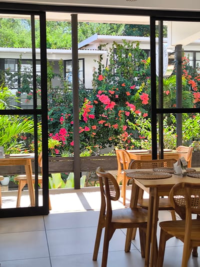 This image captures an outdoor seating area of a cafe or restaurant, likely located in the city of Goron Talo, Indonesia. The foreground features several wooden tables and chairs, set with woven placemats. In the background, vibrant pink and orange bougainvillea flowers bloom abundantly against a backdrop of green foliage and white buildings. Large glass doors and windows frame the view, suggesting a connection between the indoor and outdoor spaces. The sun casts distinct shadows on the tiled floor, indicating a bright, daytime setting. There are no people visible in the scene.