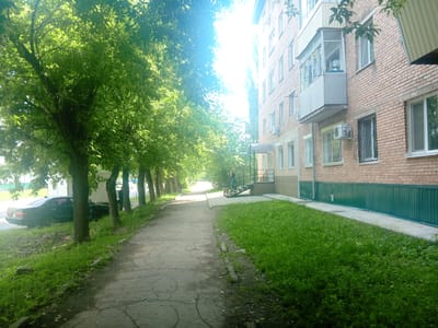 The image depicts a sunlit street scene in Arseniev, Russia. A paved walkway runs along the right side of the frame, bordered by a lush, green grassy area. On the right, a multi-story brick building with several windows and balconies is visible. The building has a green metal fence along its base. On the left side of the walkway, a line of trees with vibrant green leaves creates a canopy, casting shadows onto the path. A black sedan is parked on the left side of the street, partially obscured by trees. In the distance, a person can be seen standing at the top of an outdoor staircase leading to the building. The sun is bright, creating lens flare and washing out parts of the sky and trees.