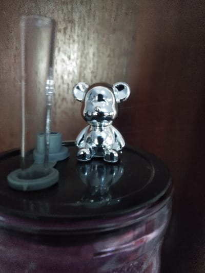 The image depicts a still-life arrangement situated in Linkebeek, Belgium. Centered on a glossy, dark-colored circular lid, a small, highly reflective chrome or silver-colored bear-shaped figurine stands upright, facing forward. To the left of the figurine, a clear, slender cylindrical plastic or glass tube, consistent with a fermentation airlock or hydrometer, is inserted into a grey plastic base that also rests on the lid. The lid itself covers a large, clear glass container, the upper portion of which is visible, revealing purple-hued contents inside. The background consists of a uniformly dark brown, textured surface, possibly wood or a wall, exhibiting subtle reflections. No people are present, and the objects are static.