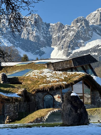 The image displays a unique, multi-level, earth-sheltered building in Going am Wilden Kaiser, Austria, situated against a backdrop of towering, snow-covered mountains under a clear blue sky. The structure features roofs primarily covered with dry, yellowish-green grass and irregular patches of white snow. Several rectangular skylights are integrated into these green roofs. The building's facades are constructed from natural, varied-sized stones. Prominent architectural elements include large modern glass panels, some triangular and some rectangular, and at least one rounded arched entryway or window on a lower level. An upper section of the building exhibits a steeply angled roof clad in wooden shingles. In the foreground, the ground is covered with a thin layer of snow, interspersed with patches of green grass. A large, dark, irregular boulder stands prominently on the snow in the lower right, with a small accumulation of snow on its peak. Bare, dark tree branches partially obscure the upper left portion of the mountain range. Faint wisps of steam or mist are visible rising from the ground level near the building's base in the lower right. No individuals or active interactions are depicted.