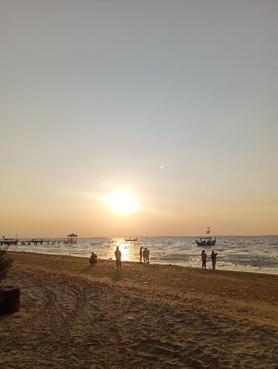 The scene depicts a beach in Tritunggal, Indonesia, during sunset or late afternoon. The sun, a bright luminous disc, is low on the horizon, centered horizontally, and reflecting intensely on the calm sea. A small white moon is visible in the pale blue-grey sky above the sun's glare, positioned in the upper right quadrant. The immediate foreground consists of sandy beach displaying distinct tire tracks and footprints. A wooden pier with a small, covered structure at its end extends from the left side into the water, with several small boats moored in its vicinity. A larger, traditional boat, featuring an upright flag, is present in the mid-right section of the water. Approximately ten human figures are distributed across the scene: one figure crouches on the sand in the mid-left; another stands further back near the center; and a cluster of approximately six figures stands near the water's edge on the right, some appearing to observe the horizon or interact with each other. All human figures are partially or fully silhouetted against the bright backlight.