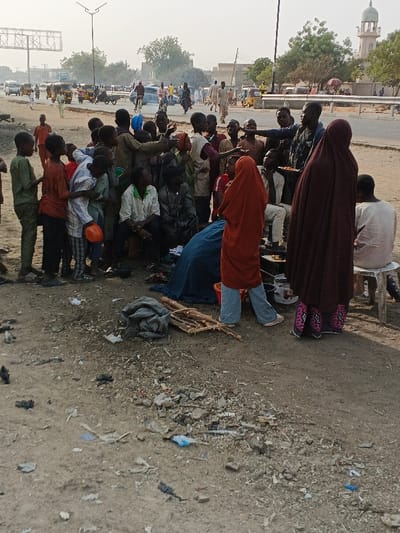 The image captures an outdoor scene in Gobirawa, Nigeria, on dusty, unpaved ground adjacent to a paved road. Approximately 20-25 individuals are visible, primarily children and young males, with several adults.

In the midground, a dense crowd of mostly children and youths gathers around a central point of interaction. A young male in a dark shirt holds a plate with what appears to be food, extending it towards the surrounding group, many of whom have hands outstretched, indicating receipt or anticipation. Another individual near him is also actively engaging with the crowd. To the right, two women are positioned: one wears a red head covering and light blue trousers, standing near a small pot on the ground; the other wears a dark brown full-length garment with pink patterned footwear, her back mostly to the viewer. A man in a white tunic sits on a low white stool on the far right. A blue tarp or fabric is spread on the ground in the center, and a bundle of dark cloth with a pile of wooden sticks or firewood lies on the dusty surface in the lower-middle foreground. The ground is scattered with small debris, including plastic pieces and refuse.

The background features a busy paved street with various vehicles, including cars, motorcycles, and auto-rickshaws, along with pedestrians. Buildings and trees line the thoroughfare, and an overhead metal sign structure is visible on the left. A light-colored mosque with a prominent minaret stands in the far-right background. The sky is bright and slightly hazy, suggesting daylight conditions.