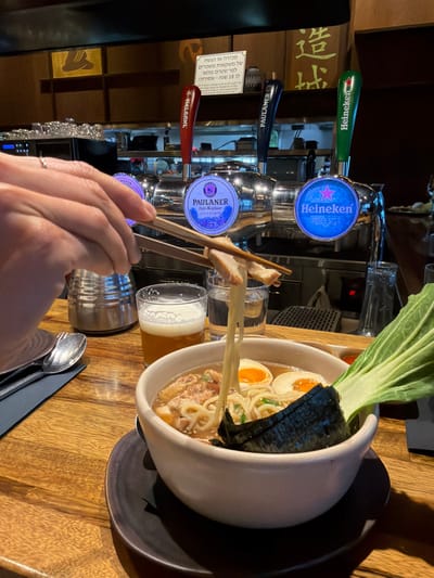 The image depicts an indoor dining scene at a restaurant or bar in Pardes Hana - Karkur, Israel, focusing on a meal and beverages. In the immediate foreground, a person's hands are visible holding wooden chopsticks, lifting a strand of ramen noodles and a piece of meat from a white ceramic bowl. The ramen bowl, resting on a dark saucer on a wooden table, contains broth, wavy noodles, two halved soft-boiled eggs, chopped green onions, sliced meat, a large piece of nori, and a vibrant green bok choy leaf. To the left of the ramen bowl, a metal spoon and fork are placed on a dark grey napkin. Behind the ramen, a clear glass filled with a light amber, foamy beverage (likely beer) is visible, alongside another clear glass containing water. Further left, a cylindrical silver metal container stands on the table.

The midground is dominated by a polished chrome draft beer system with three distinct tap handles. From left to right: a red tap handle with a star logo (partially identifying "Goldstar"), a dark blue tap handle clearly labeled "PAULANER" with a round illuminated medallion beneath it displaying "PAULANER Hefe-Weißbier NATURTRÜB", and a green tap handle clearly labeled "Heineken" with an illuminated medallion showing the Heineken logo.

In the background, behind the beer taps, various stainless steel bar equipment is visible. Mounted on a wooden wall, a white sign with Hebrew text reads: "מכירה או הגשה של משקאות משכרים למי שטרם מלאו לו 18 שנה - אסורה!" (Sale or serving of alcoholic beverages to anyone under the age of 18 - forbidden!). To the right of this sign, Japanese characters "造城" (Zàochéng) are visible as part of the wall decor. The overall environment is dimly lit with a warm ambiance.