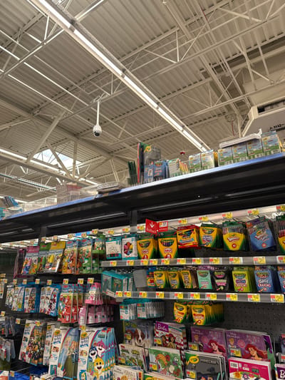 This image captures a rotated view inside a retail store, likely a discount department store in Worcester, United States. The scene primarily focuses on an aisle displaying children's art supplies and activity books, with a significant portion of the store's ceiling also visible.

On the left side of the rotated image (which would be the top in standard orientation), the store's high, industrial-style ceiling is prominent. It features a network of white metal trusses, exposed air ducts, and long rows of fluorescent light fixtures, providing bright illumination. A white security camera is mounted from the ceiling structure, monitoring the aisle.

The right side of the rotated image showcases store shelves filled with a wide array of products. These dark-colored shelves are stocked with numerous items geared towards children's creativity. Visible products include various boxes of "Crayola" crayons, such as "JUMBO" and "NEON CRAYONS," as well as activity books featuring popular franchises like Disney "PRINCESS," "MINECRAFT," and "Barbie." Other items like "MESS FREE" activity kits and "PET PALS" products are also displayed. Price tags are visible on many items, along with product count numbers (e.g., "24," "16," "12," "10," "48"). A red "Rollback" sign with an arrow pointing to the left/down is also visible on one of the shelves, a common feature in stores like Walmart. Partially visible text on an overhead sign reads "AHU-" along with isolated letters "B" and "S." The overall scene suggests a busy and well-stocked shopping environment.