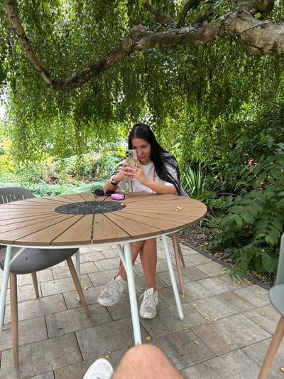 A young woman is sitting at a round, wooden outdoor table, holding a smartphone and looking at its screen. She has long, dark hair and is wearing a white t-shirt and light-colored sneakers. On the table, next to her, are a pair of sunglasses and a small, purple object. The table is set on a patio paved with rectangular stone tiles. Behind her, a lush green tree with hanging branches provides shade. In the background, there is dense vegetation. The scene is an outdoor seating area, likely a garden or patio, located in Koszuty-Huby, Poland. The overall atmosphere is relaxed and natural.