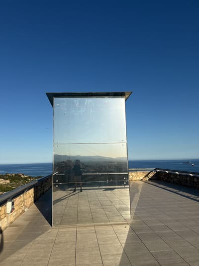The image shows a rooftop observation point on a sunny day in Tarragona, Spain. A three-paneled mirror structure stands on the tiled rooftop. The reflection in the mirror shows a person taking a photo of themselves, along with the cityscape and the sea in the background. A stone railing surrounds the rooftop edge, offering a view of the coastline and ocean. The sky is clear and blue. The location is likely a high vantage point within the city of Tarragona, possibly a hotel or public observation area. The overall impression is of a scenic location designed for viewing and photography.