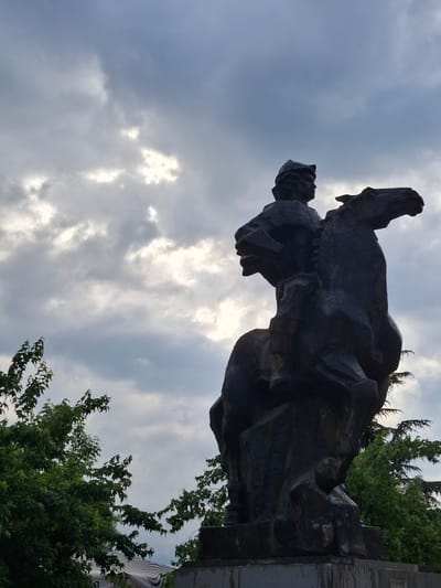 The image shows a statue in Krumovgrad, Bulgaria. The statue depicts two figures, one appearing to carry or support the other. The statue is dark in color and sits against a cloudy sky. Green trees are visible in the background on the right side of the frame.