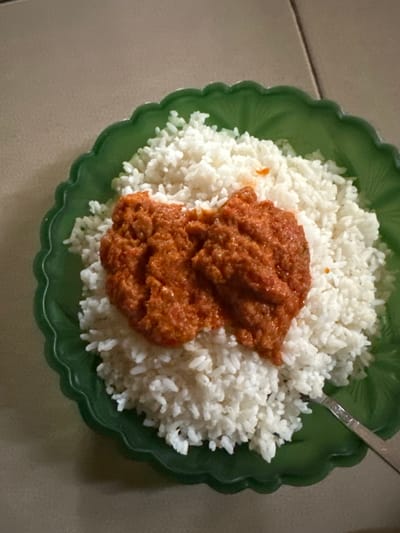 A green, scalloped plastic plate holds a serving of white rice topped with a generous portion of reddish-orange stew or sauce. A metallic spoon, partially visible, rests on the lower right edge of the plate. The plate and its contents are situated on a light-colored, possibly beige or cream-tiled surface with visible grout lines, suggesting an indoor setting. No individuals or specific actions are depicted. The scene is located in the city of Ketu Adieowe, Nigeria.