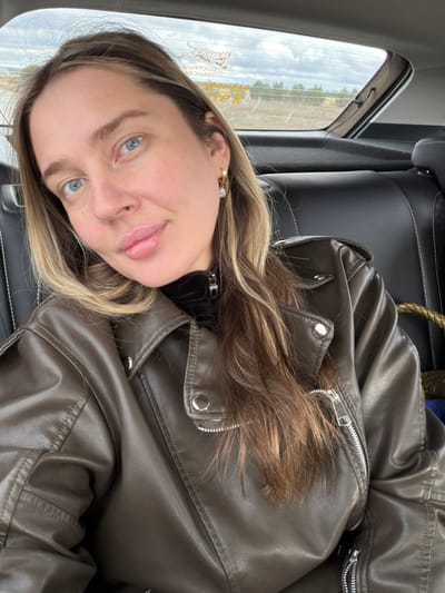 A woman is in the back seat of a car, wearing a brown leather jacket. The scene outside the window shows a field with some trees and buildings in the distance. In the window there is some text in a foreign language which is likely Russian. Based on that information, the location is likely in Povarenki, Russia.