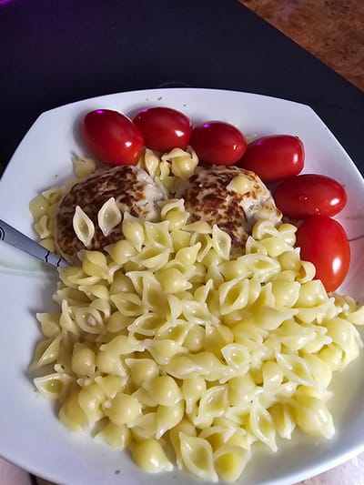 A close-up, overhead shot features a white plate of food on a dark surface. The plate contains a portion of shell pasta, two cooked hamburger patties, and several small, red cherry tomatoes. The pasta is a pale yellow and appears to be coated in a light sauce. The hamburger patties are round and speckled with brown, suggesting they are well-cooked. The cherry tomatoes are clustered to the left of the plate, with some showing a slight sheen that may be water droplets. A fork with a metallic handle is visible at the bottom right, partially submerged in the pasta. The background is out of focus, showing a dark, possibly black tablecloth, with a hint of a wood-grain pattern and purple lighting on the left side. The scene appears to be an indoor setting, likely a meal. No text is visible.