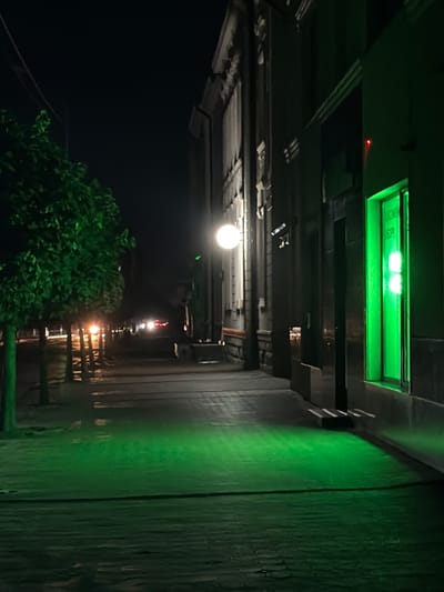 This image depicts an outdoor scene at night, possibly in Stryi, Ukraine. The perspective is from below, looking upwards and outwards.
The primary subjects are various light sources and architectural elements. A prominent feature is a bright green, rectangular light fixture mounted above, emitting a strong, artificial glow. Below this, there are several horizontal beams and structures, suggesting an overhead walkway, bridge, or building overhang. A single, round, white light hangs lower down, illuminating the immediate area. Further into the distance, a series of smaller, warmer lights are visible, receding along a pathway or road.
On the right side of the image, a textured, vertical surface, possibly a brick or concrete wall, is illuminated by a wash of green light, creating a stark contrast with the surrounding darkness. On the left, a dense, dark green tree or shrubbery is visible in the foreground, its foliage absorbing much of the ambient light.
The overall impression is one of an urban or semi-urban environment at night, likely with artificial lighting playing a significant role in shaping the scene. The presence of the green lights could indicate signage, decorative lighting, or functional illumination for a specific area. The image conveys a sense of quietness and perhaps a slightly eerie or dramatic mood due to the strong artificial lighting and dark surroundings. No people are visible in the frame. The time of day is clearly night.