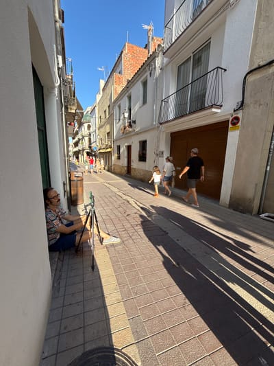 This media file captures a sunny day in a narrow European street, likely in Tossa De Mar, Spain, based on the architectural style and tiled ground. The perspective is from ground level, looking up a sloped street lined with white and brick buildings.
In the foreground, a man with glasses and a colorful short-sleeved shirt lies on the ground next to a tripod, seemingly relaxing or observing the scene. His posture suggests a casual and unhurried activity.
Further up the street, three people are walking away from the camera. The person in front is a man wearing a black t-shirt and shorts, followed by a woman in a white top and shorts, and a child in a white dress. They appear to be enjoying a leisurely stroll.
The street is paved with light-colored square tiles, and the bright sunlight casts long, sharp shadows from unseen objects, indicating it is likely midday. The sky is a clear, vibrant blue, suggesting fair weather. One of the buildings has a closed wooden garage door with a circular sign above it. The sign features red and black imagery, possibly a prohibition symbol. Another building displays an antenna on its roof. The overall impression is one of a warm, bright day in a picturesque, possibly historic, urban environment.