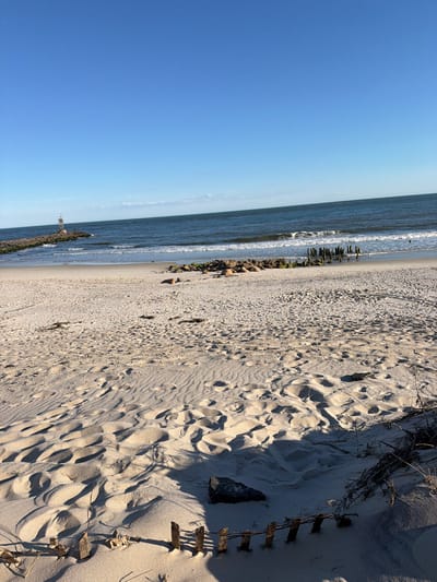 The image depicts a tranquil beach scene in Hampton Bays, United States of America. In the foreground, the sandy beach is textured with numerous footprints and shallow depressions, suggesting recent human activity. A low, weathered wooden fence, partially buried in the sand, runs along the bottom right of the frame. The sand is illuminated by the warm glow of what appears to be late afternoon sunlight, casting long shadows.
Further back, the gentle waves of the ocean meet the shore, creating a foamy white surf. In the distance, a stone jetty extends into the water, topped by a small white structure with a lantern, likely a lighthouse or navigational marker. Several wooden pilings, remnants of a past structure, emerge from the water near the center of the frame, adding a touch of history to the scene. The vast expanse of the ocean stretches to the horizon, meeting a clear blue sky dotted with a few wispy clouds. The overall atmosphere is serene and natural.
