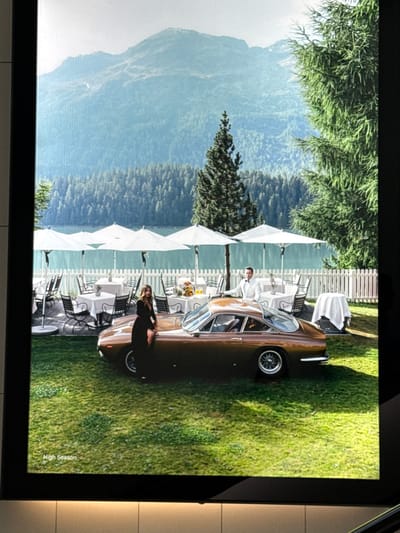 The image depicts a scene at a lakeside resort with a mountain backdrop. In the foreground, a vintage brown convertible car is parked on a manicured lawn. A woman in a black dress is leaning against the car. Behind the car, there are several white umbrellas shading outdoor dining tables, a white picket fence, and a body of water, likely a lake, with a forested hillside beyond. Further in the background, a mountain range is visible under a hazy sky. The overall impression is one of luxury and leisure. The text "High Season" is visible in the bottom left corner of the image, suggesting a promotional or artistic context. The scene is not located in Sesto San Giovanni, Italy, as this is an outdoor scenic view typical of alpine regions.