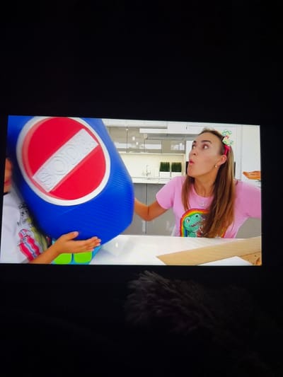 The image shows a woman wearing a pink t-shirt with a dinosaur graphic and a colorful bow in her hair, looking surprised. She is in a room that appears to be a kitchen, with modern cabinets and countertops visible in the background. In the foreground, a large blue object with a red circle and a white diagonal stripe, resembling a "no soda" sign, is being held by a child whose hands are visible. The child is wearing a white t-shirt with colorful stripes. The overall scene appears to be from a video recording, possibly a children's educational program or a vlog. There is no information in the image to suggest any location in Tambov, Russia.