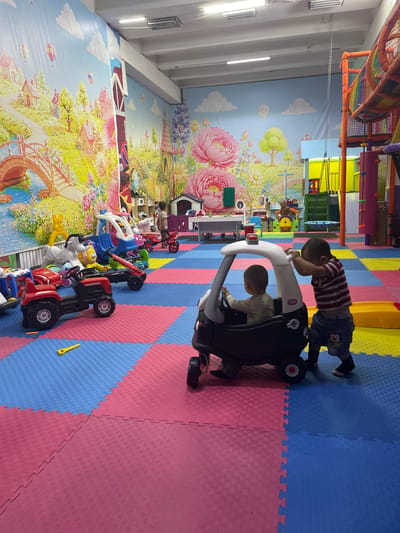 In a brightly lit indoor playground with a colorful, cartoon-themed mural depicting a whimsical landscape of houses, bridges, and hot air balloons, two young boys are engaged in play. The floor is covered with interlocking foam mats in shades of blue, pink, and yellow.

The primary subjects are the two boys. One boy, wearing a striped t-shirt and shorts with a cartoon character, is standing and leaning over a black toy car, appearing to push it. Another boy, dressed in a light-colored shirt, is seated inside the same toy car, holding onto its steering wheel. The toy car is a small, ride-on vehicle with a white steering wheel and a black body.

The scene is set in what appears to be a children's play area or a soft play center. The vibrant murals and the presence of multiple toy vehicles, including a red tractor and a blue ride-on toy, suggest a playful and stimulating environment designed for young children. There are no clear indicators of the time of day or weather conditions as it is an indoor setting. The emotions of the children are not explicitly discernible, but their engagement with the toys suggests they are enjoying themselves.

The overall atmosphere is one of active play and childhood fun. The location context of Batken, Kyrgyzstan, is provided, but there are no specific visual cues within the image that would exclusively identify it as such. No visible text is present in the image, apart from what might be branding on the toy cars, which is not legible.