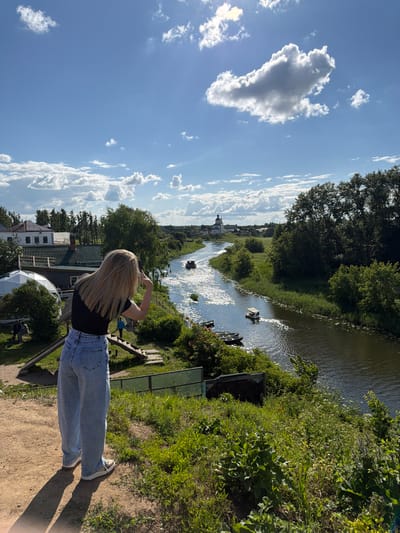 The image depicts a scenic view of a river in Suzdal, Russia, on a sunny day with scattered clouds. In the foreground, a young woman with blonde hair stands with her back to the camera, wearing a black t-shirt and light-wash wide-leg jeans. She is looking out towards the river and holding up two fingers in a peace sign. To her left, there are some buildings with green roofs and a white domed structure. The river flows through a green, tree-lined landscape, and several small boats are visible on its surface. In the distance, a white church with a green dome stands prominently against the sky. The overall scene conveys a sense of tranquility and natural beauty.