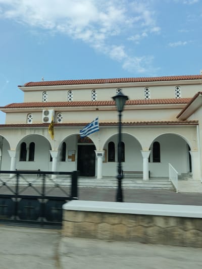 A side view of a tan-colored church with a red tiled roof is shown under a blue sky with scattered white clouds. The church features a series of arched entrances supported by white columns. Above the arches, there are rows of small, decorative windows with a cross-like pattern. A Greek flag and a yellow flag are visible on poles outside the church. A black lamppost stands in front of the building. In the foreground, a black metal fence is partially visible, with a stone wall and a paved area behind it, suggesting the church is located on a street. The image appears to be taken from a moving vehicle due to slight motion blur. The location is identified as the Municipal Unit of Sikyona, Greece.