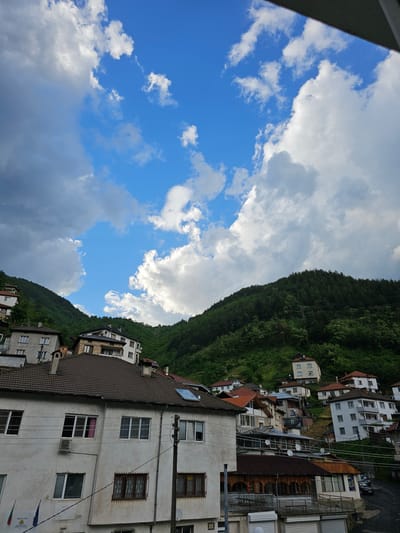 The image shows an urban landscape in Erma Reka, Bulgaria. A densely vegetated hillside rises behind closely packed buildings, with a clear blue sky interspersed with white and gray clouds above. The buildings are a mix of residential and commercial, displaying a variety of architectural styles and colors. Power lines are visible, crisscrossing between structures.