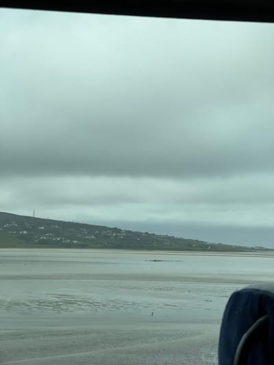 This media file depicts a landscape scene. The dominant subjects are a vast expanse of wet sand, likely a beach at low tide, and a distant, overcast sky. A green, hilly terrain with scattered buildings is visible along the left side of the frame, suggesting a coastal village or town. The sand has parallel, elongated textures, possibly from receding water or wind patterns.

There are no clearly discernible people or specific objects that stand out as the primary focus, though there are some small dark shapes on the sand that could be distant figures or debris. A dark blue object, possibly a piece of clothing or equipment, is visible in the upper right corner.

The weather appears to be overcast and possibly humid, contributing to the muted color palette of the scene. The time of day is indeterminate due to the lack of direct sunlight and shadows. The overall mood is calm and somewhat somber, due to the gray skies.

The location context provided is Killult Middle, Ireland. This information, combined with the visual cues of the coastline and the architecture, helps to place the scene. There is no visible text in the image. The image is oriented vertically, with the top of the frame being the left side in a typical horizontal view.